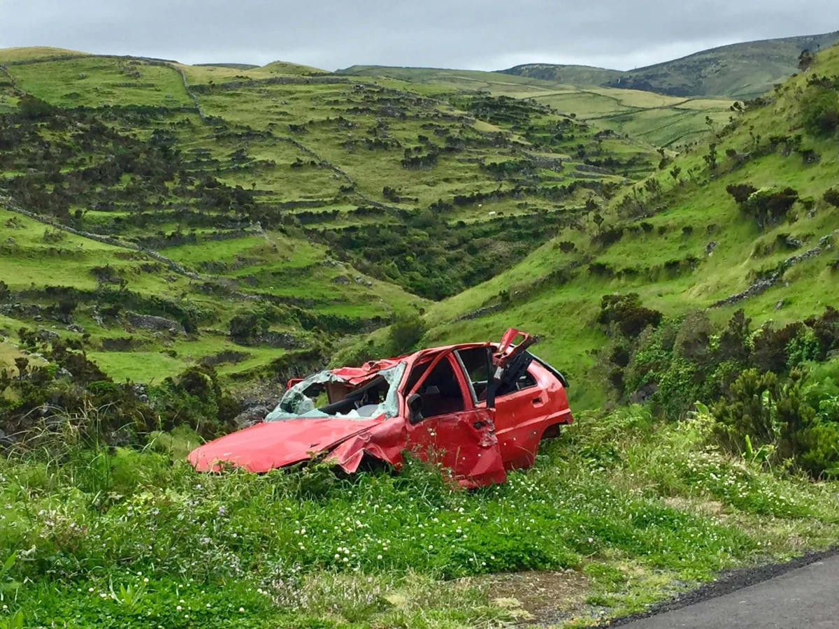 A heavily damaged red car abandoned on a grassy slope against green terraced hills.