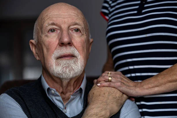 Elderly man looking thoughtful as a caregiver places a supportive hand on his shoulder.