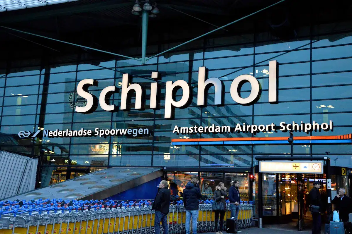 Glass facade of Amsterdam Airport Schiphol featuring large signage and luggage carts in front.