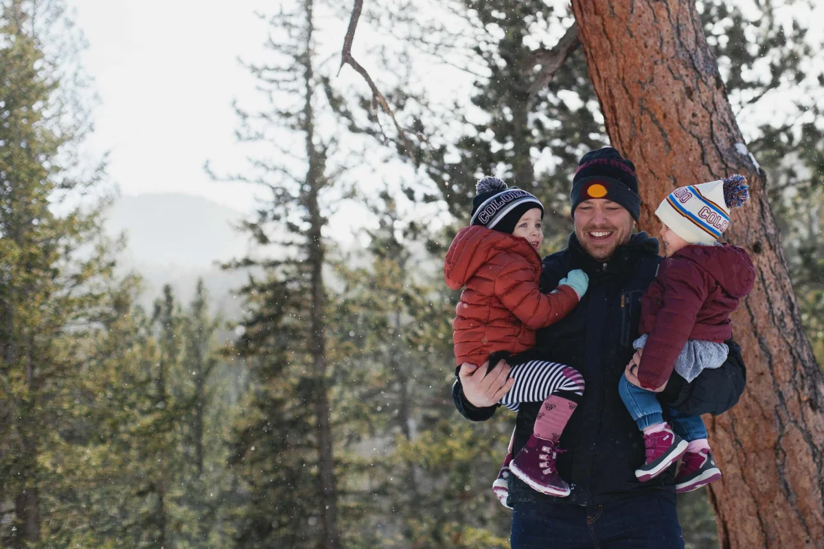 A smiling father holds two young children wearing winter clothes in a snowy forest.