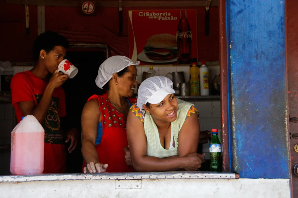 Two women wearing white hairnets smile inside a food stall while a man drinks.