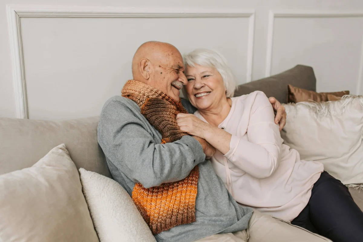 An elderly couple smiling and embracing while sitting together on a comfortable couch.