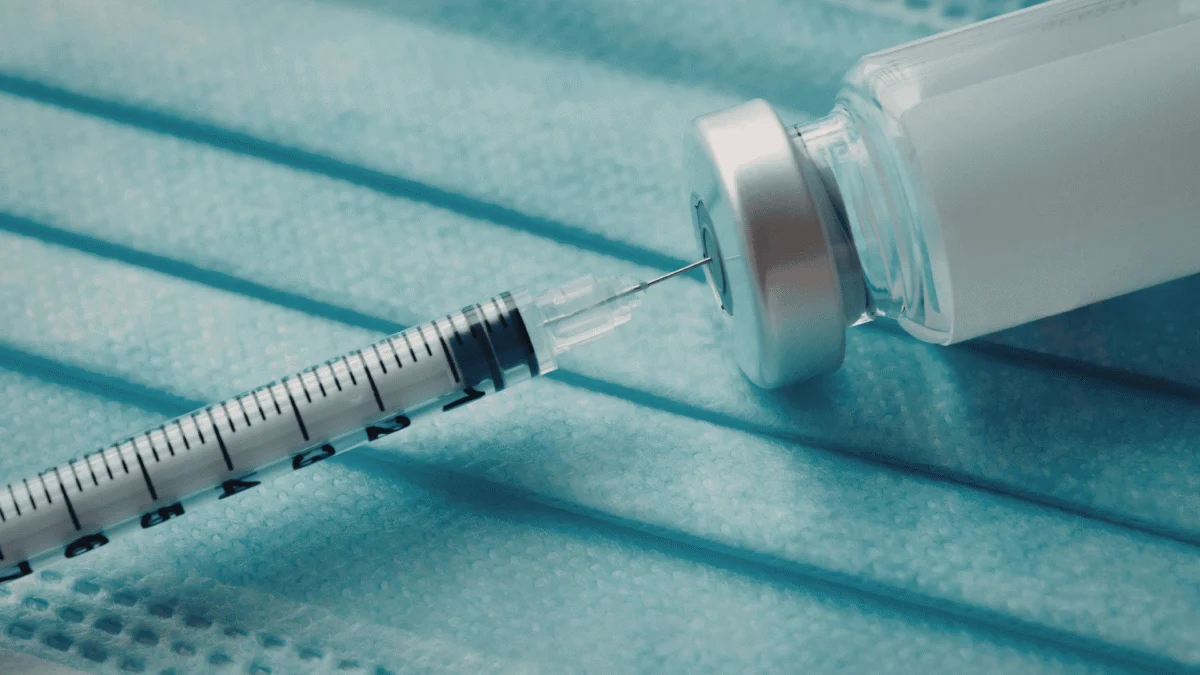 Syringe needle inserted into a vaccine vial lying on a blue surgical mask