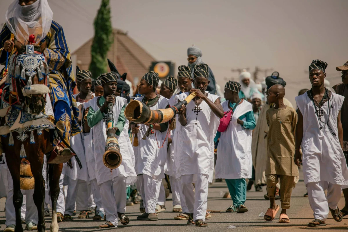 Men in traditional white attire playing long horns during a vibrant street procession.