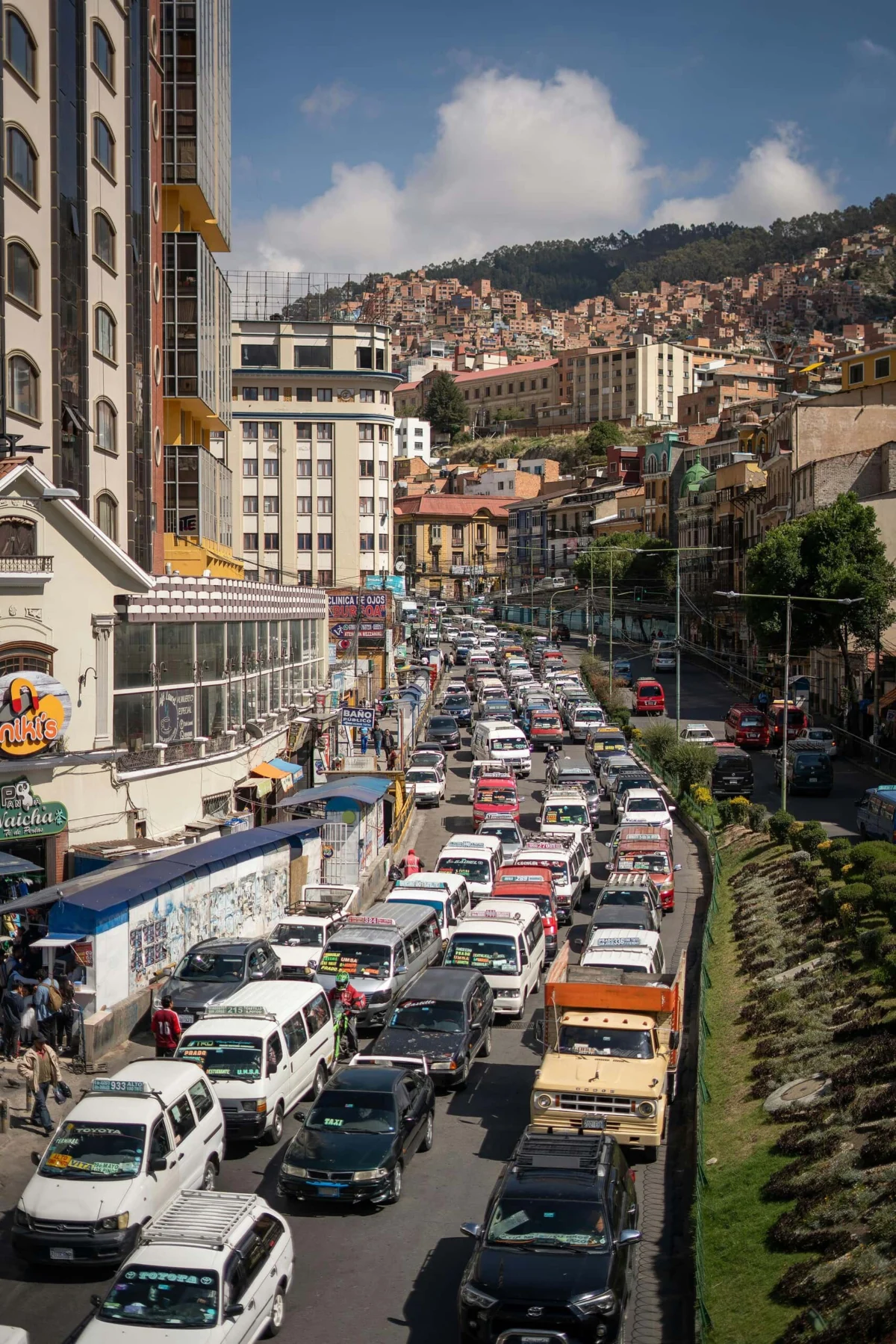 Busy city street packed with cars and minibuses with dense hillside buildings in the background.