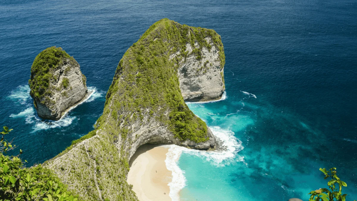 Green cliff formation surrounding a sandy beach in vibrant blue and turquoise ocean water.