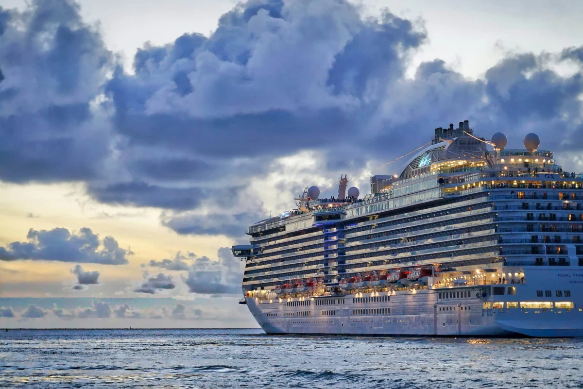 Large Regal Princess cruise ship sailing on ocean waters under a dramatic cloudy sunset sky.