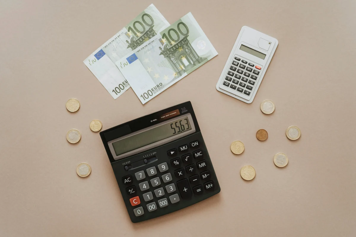 Flat lay of two calculators, Euro banknotes, and coins scattered on a beige surface.