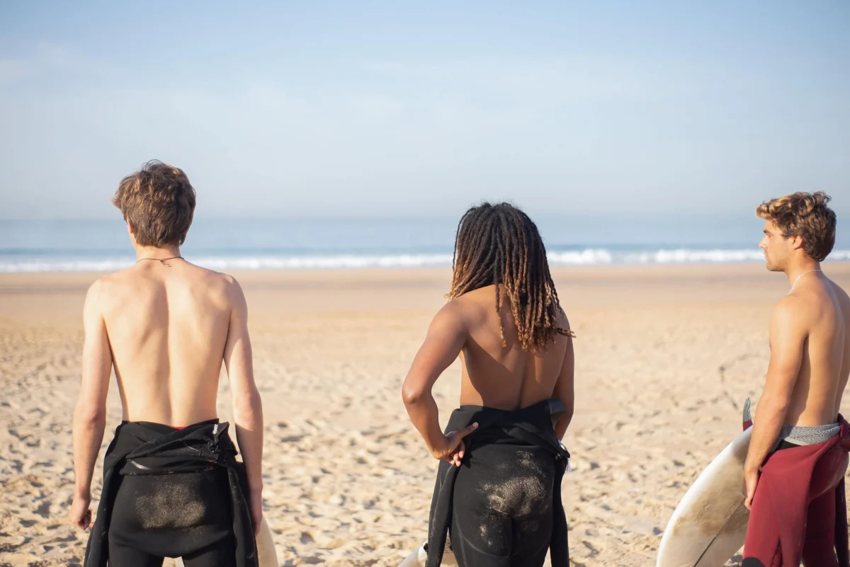 Three young surfers standing on a sandy beach looking out towards the ocean.
