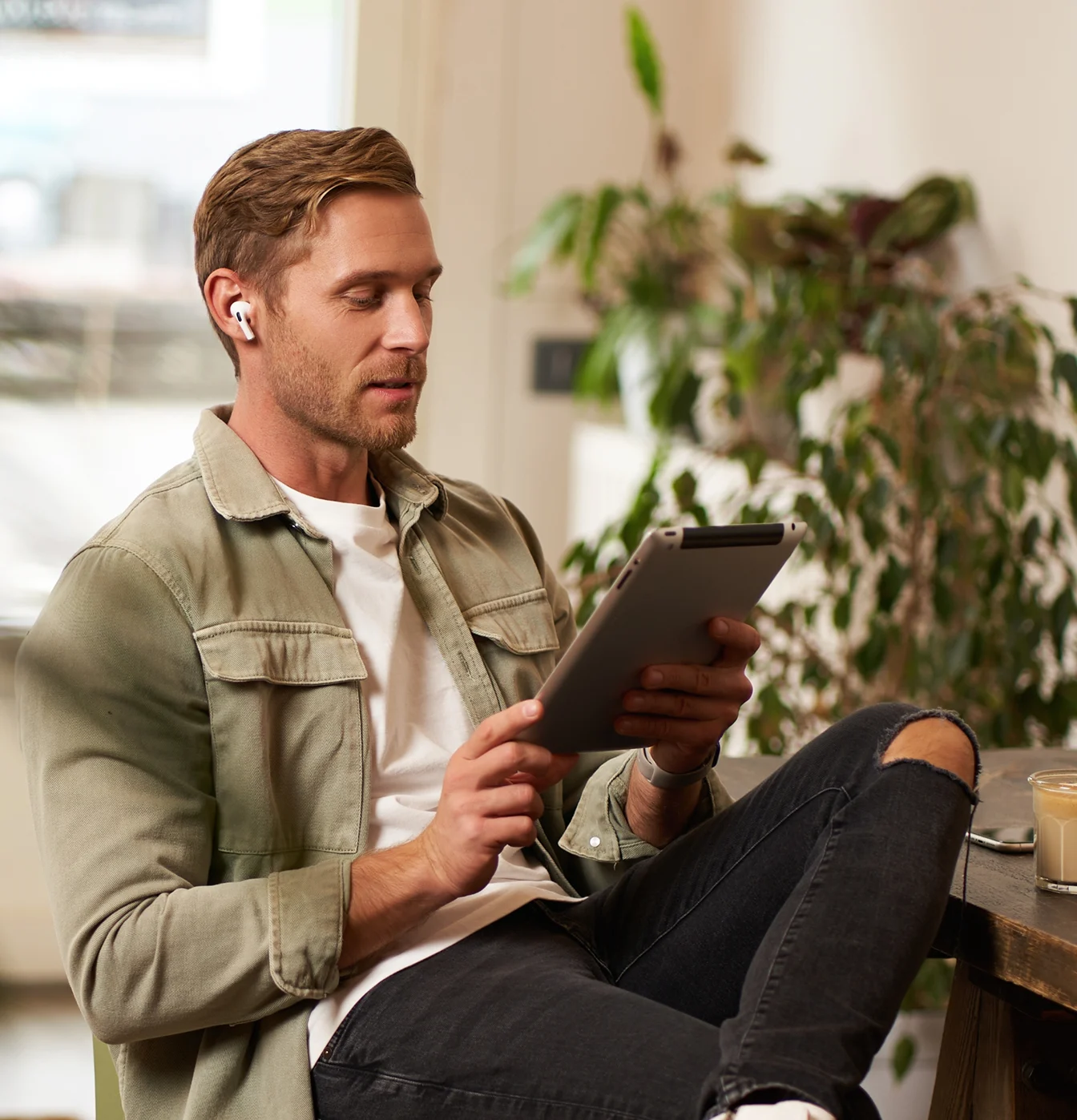 A man wearing wireless earbuds sits at a table using a digital tablet.