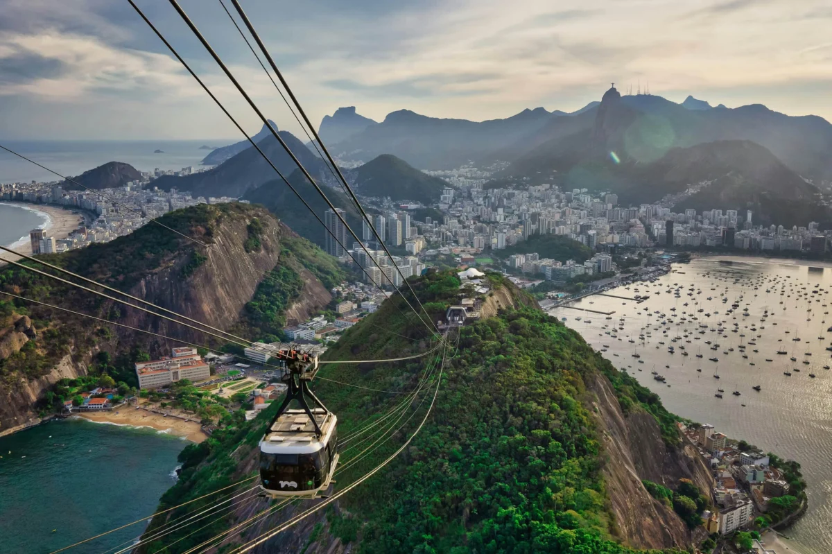Cable car riding above green hills with Rio de Janeiro coastline and mountains in background.