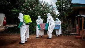 Four health workers in full white hazmat suits carrying supply boxes outdoors.
