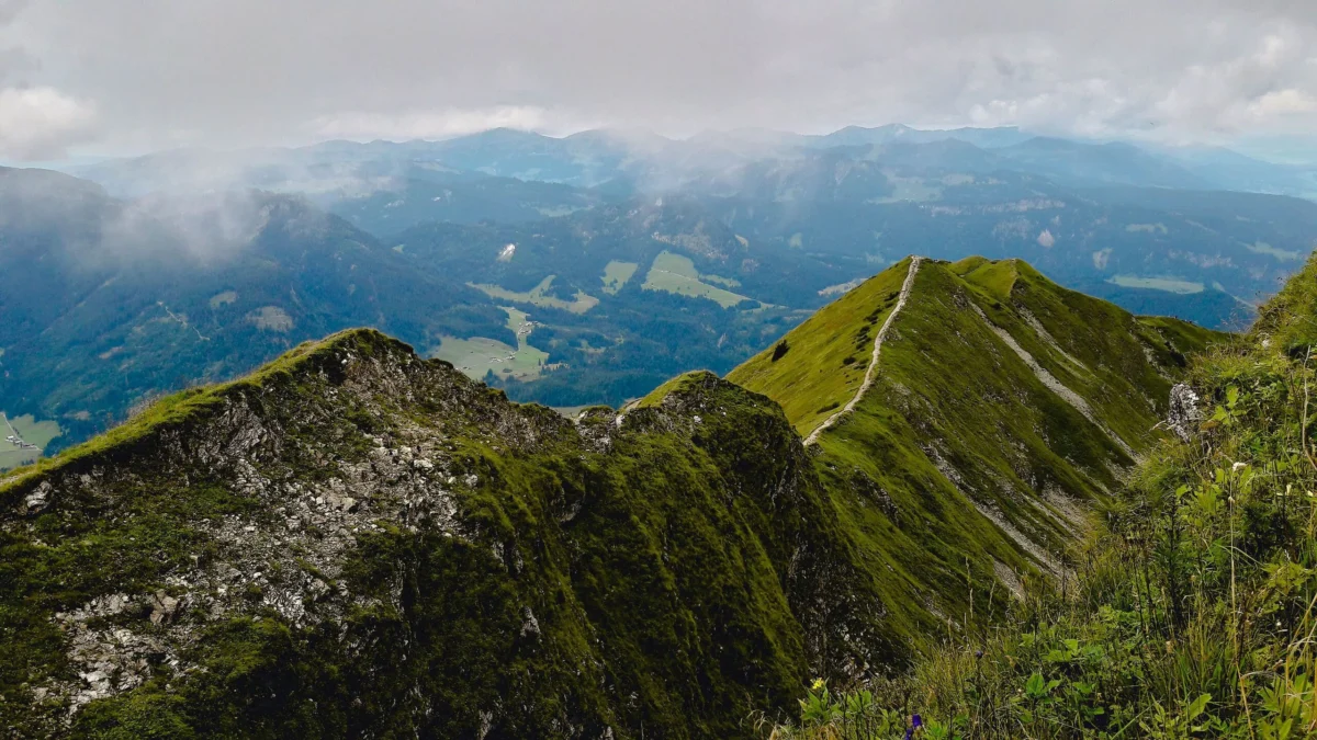 Narrow trail running along a steep green mountain ridge under a cloudy sky.