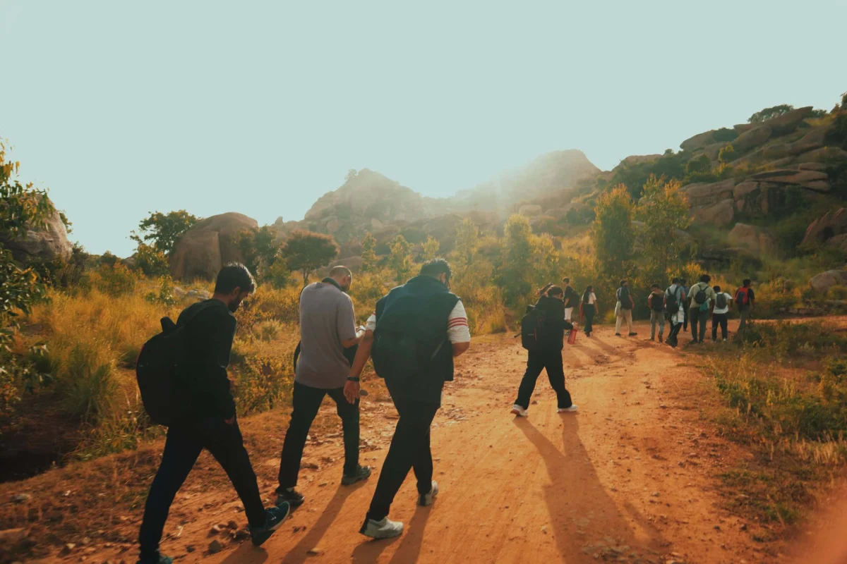 A group of people with backpacks hiking along a sunny dirt path through rocky hills.