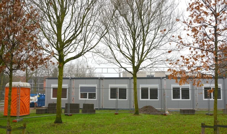 Modular grey office cabins behind a construction fence on a grassy lawn with trees.