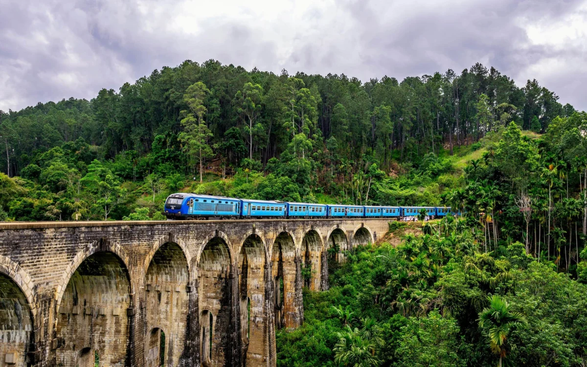 Blue passenger train crossing the historic Nine Arch Bridge surrounded by lush green forest