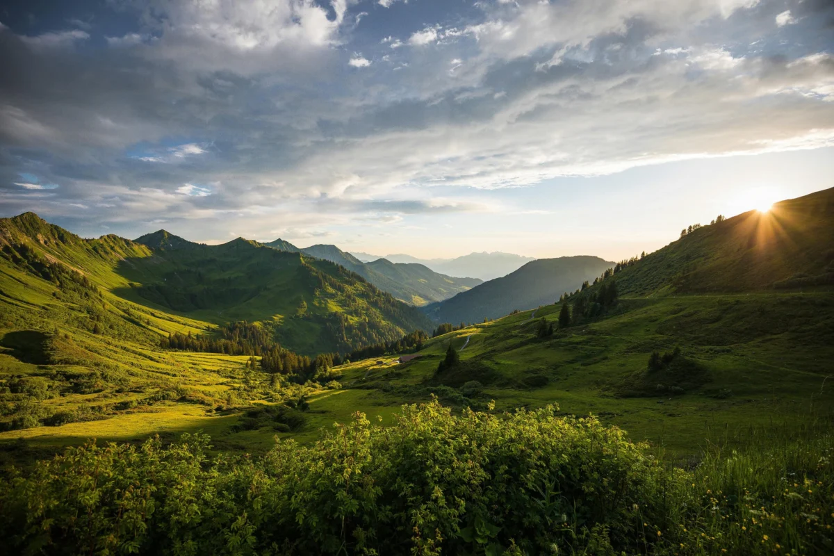 Sunlight peeking over lush green mountain slopes and a grassy valley under cloudy skies.