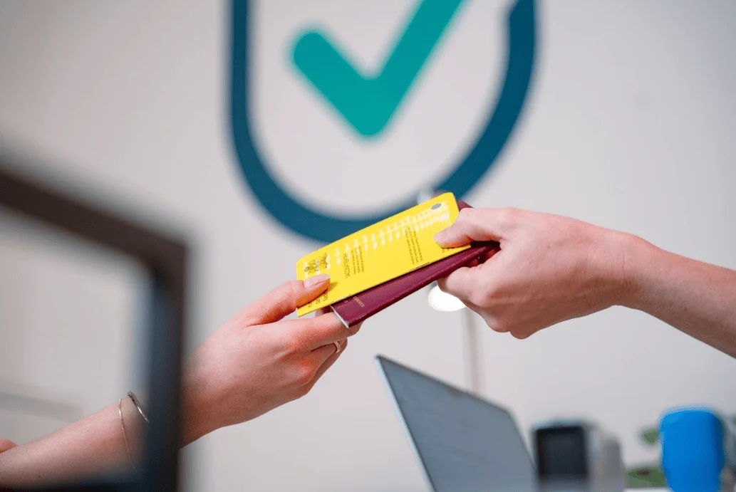 Two hands exchanging a passport and yellow vaccination certificate at an office counter.