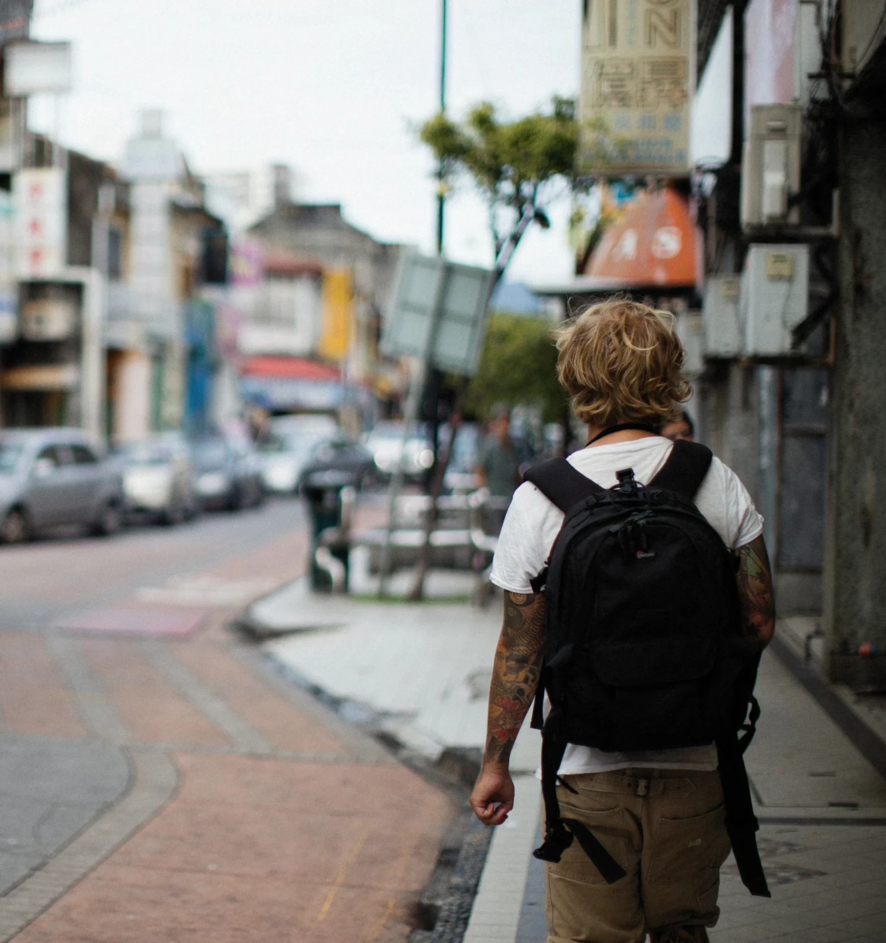 Person with a black backpack walking down an urban street lined with parked cars.