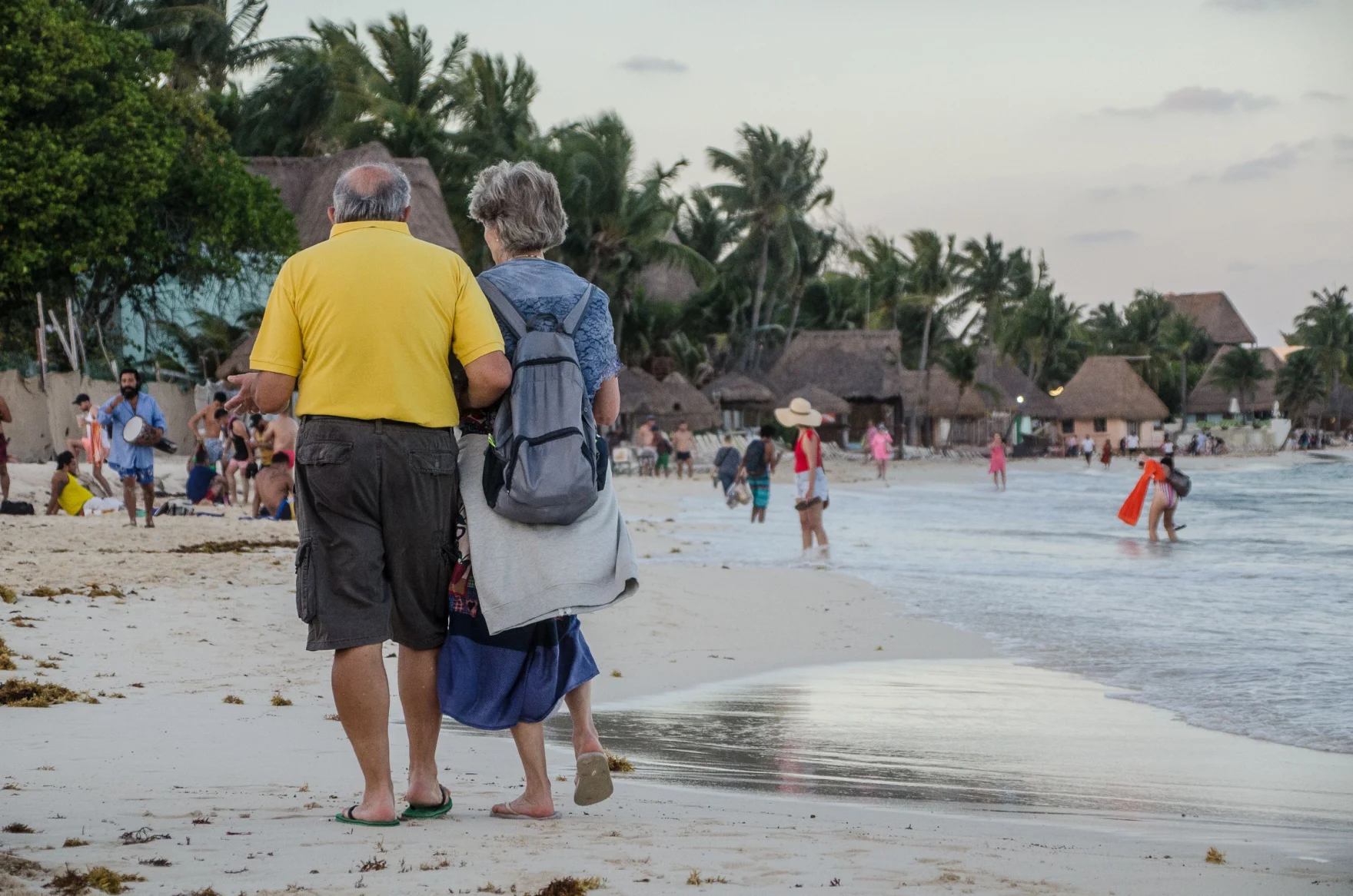 Senior couple walking along a sandy tropical beach with palm trees and huts