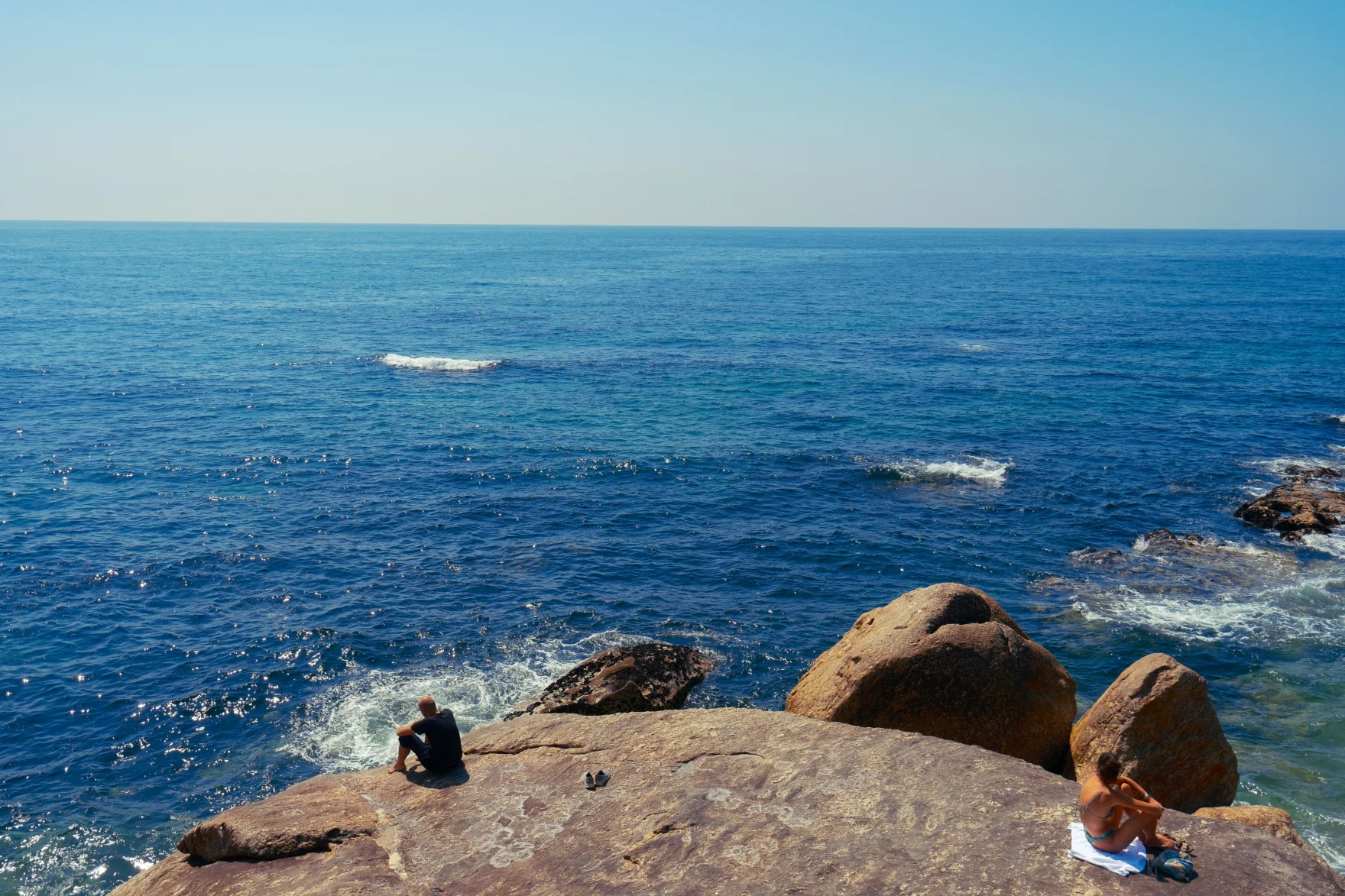 Two people relaxing on rocky coastal boulders overlooking the vast blue sea.
