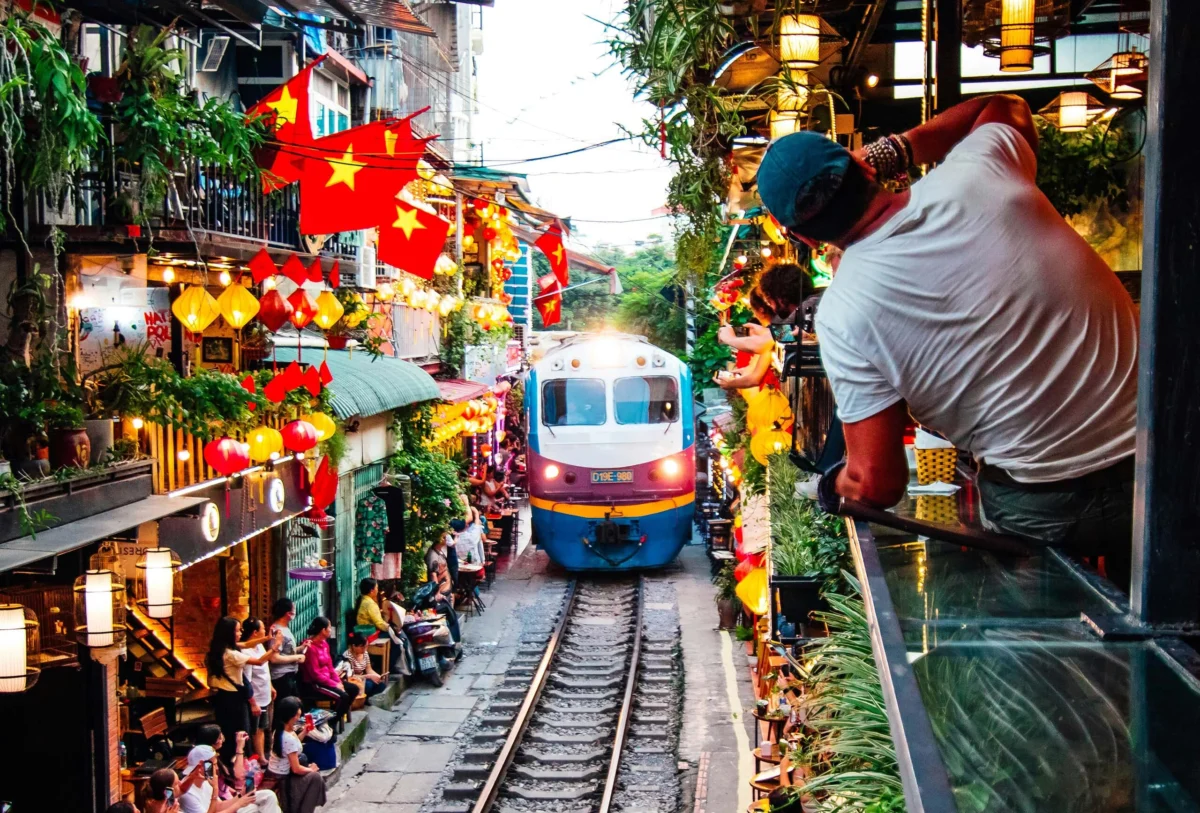 Train passing through narrow Hanoi Train Street lined with cafes, lanterns, and Vietnamese flags